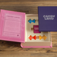 The Candy Land Vintage Bookshelf Edition by WS Game Company is shown opened, revealing the game pieces and instructions contained within a pink and purple box on a wooden surface—ideal as a child's first game or as a collectible edition.