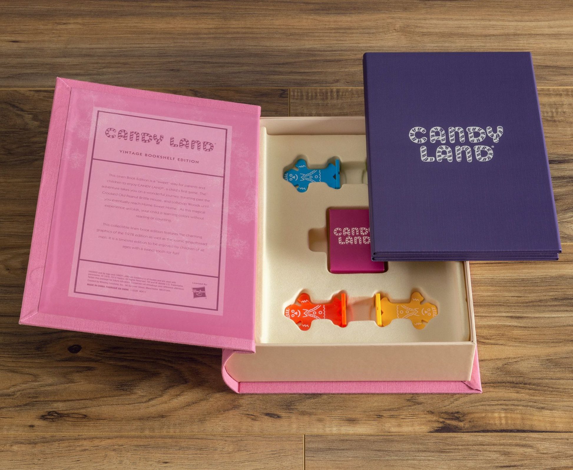 The Candy Land Vintage Bookshelf Edition by WS Game Company is shown opened, revealing the game pieces and instructions contained within a pink and purple box on a wooden surface—ideal as a child's first game or as a collectible edition.