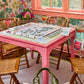 An Oh My Mahjong Mat Collection pink table is set with a board game, surrounded by bamboo chairs. Colorful floral-patterned wallpaper and a window complete the scene, with a tote bag visible in the foreground.