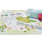 The Oh My Mahjong Mat Collection by Oh My Mahjong features a pastel green patterned neoprene non-slip mat, card racks, rule cards, and a floral bag. The set also includes mahjong tiles scattered across the mat.