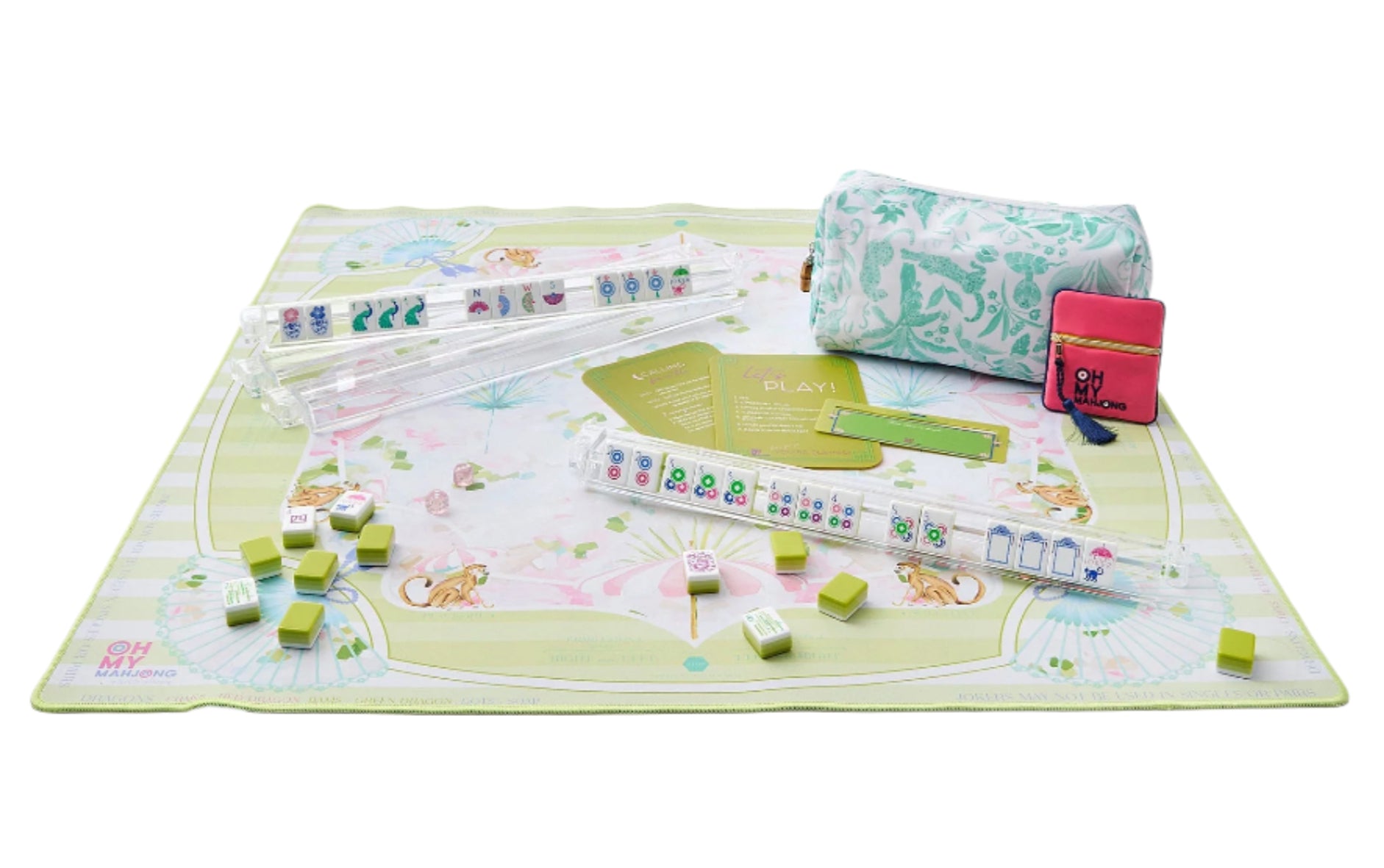 The Oh My Mahjong Mat Collection by Oh My Mahjong features a pastel green patterned neoprene non-slip mat, card racks, rule cards, and a floral bag. The set also includes mahjong tiles scattered across the mat.
