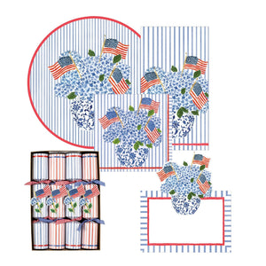 Caspari’s Flags And Hydrangeas Collection offers patriotic paper goods—placemats, napkins, crackers, and a notecard—featuring blue floral vases with flags, striped backgrounds, and red borders for festive outdoor tables.