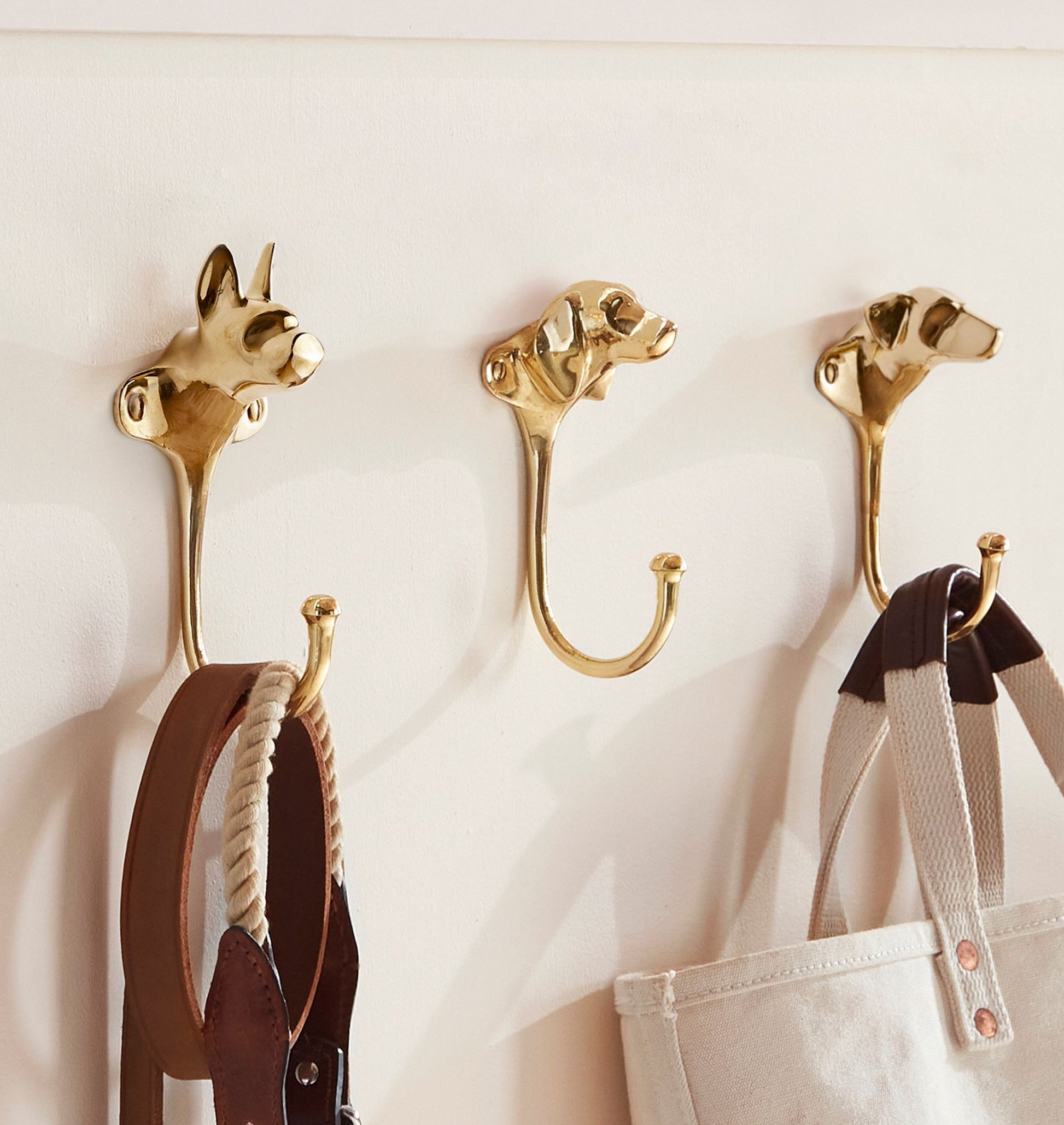 Three Cast Brass Dog Hooks by Rejuvenation, shaped like dog heads, hold a belt, a tote bag, and a leather strap against a white wall.
