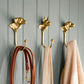 Three Rejuvenation Cast Brass Dog Hooks are mounted on gray paneling, each holding a brown leash, a light scarf, and a beige knitted sweater.
