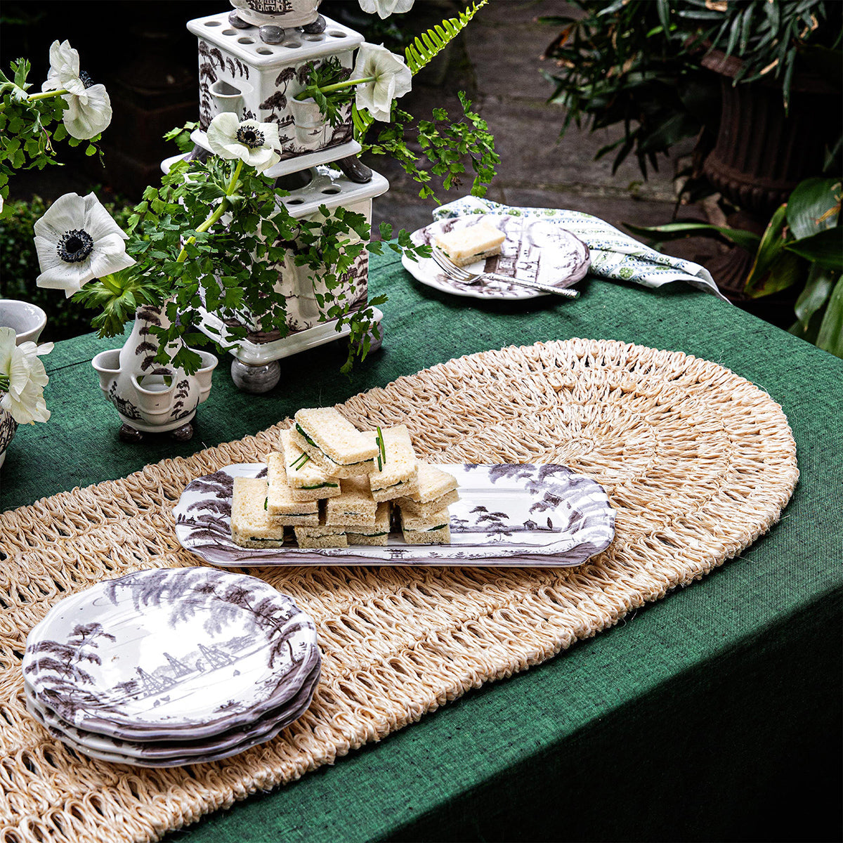 A green tablecloth with Juliska's Straw Loop Natural Table Runner, black and white china, sandwiches, napkins, and white flowers create an elegant outdoor tablescape.