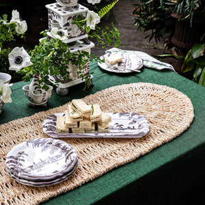 A green tablecloth with Juliska's Straw Loop Natural Table Runner, black and white china, sandwiches, napkins, and white flowers create an elegant outdoor tablescape.