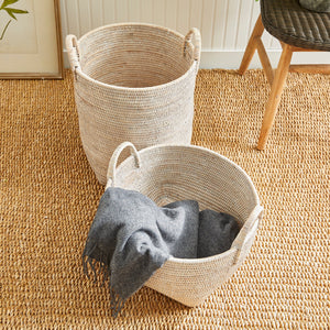 Two Burma Whitewash Rattan Orchard Baskets by Napa Home & Garden sit on a textured rug; one is empty and the other holds a folded gray blanket—ideal for organizing your space.