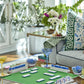 The Oh My Rummi Set by Oh My Mahjong features rummi tiles and cards on a green mat, surrounded by drinks and snacks in a modern living room—ideal for lively family game nights.