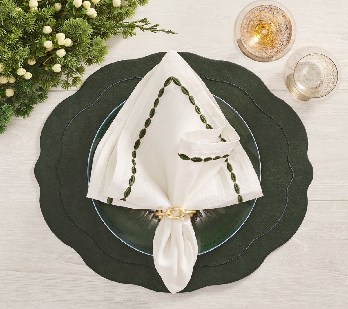 A festive table setting featuring a green charger, white plate, and an embroidered napkin elegantly secured with a Kim Seybert Chain Link Napkin Ring, accompanied by two glasses and lush greenery on a light wooden table.