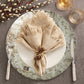 A table setting featuring the Kim Seybert Chalet Placemat, a white plate, fork and knife, beige fringed napkin tied with twine and pinecones, two glasses, and decorative pussy willow branches on a grey surface.
