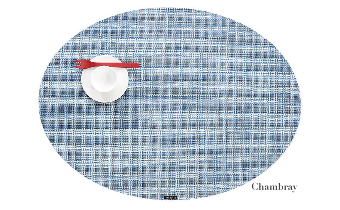A blue Oval Placemat by Chilewich, styled with a white cup and saucer and a red fork—ideal for any dining setting. "Chambray" is seen in the lower right corner.