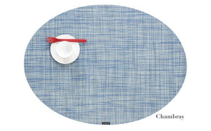 A blue Oval Placemat by Chilewich, styled with a white cup and saucer and a red fork—ideal for any dining setting. "Chambray" is seen in the lower right corner.