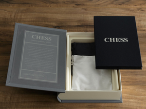 The image displays an opened Chess Vintage Bookshelf Edition box from WS Game Company on a wooden table. This luxurious edition includes a black and white cloth bag and a booklet labeled "CHESS." Text is visible on the inside cover of the box lid, making it perfect for enthusiasts of this timeless strategy board game.