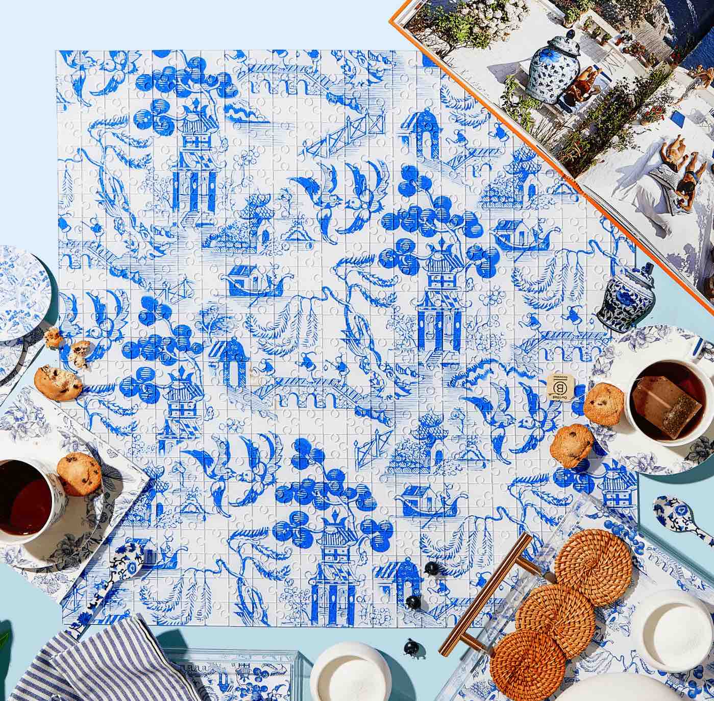 A finished Tart by Taylor Acrylic Puzzle from the Bubbly Butterfly collection, featuring blue and white floral and architectural patterns, is set on a light blue surface with tea, cookies, plates, napkins, and an open book—a charming home decor accent.
