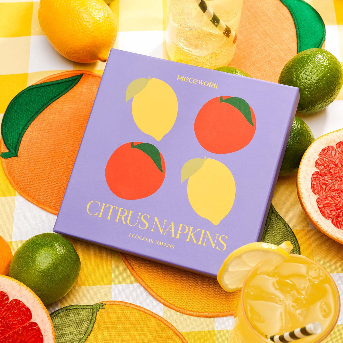 Piecework Citrus Cocktail Napkins in a square purple box, surrounded by fresh lemons, limes, grapefruits, and two glasses of citrus drinks on a yellow gingham tablecloth.