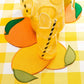 Round yellow and orange Piecework Citrus Cocktail Napkins on a yellow gingham tablecloth, set with a glass of lemonade and a pitcher.