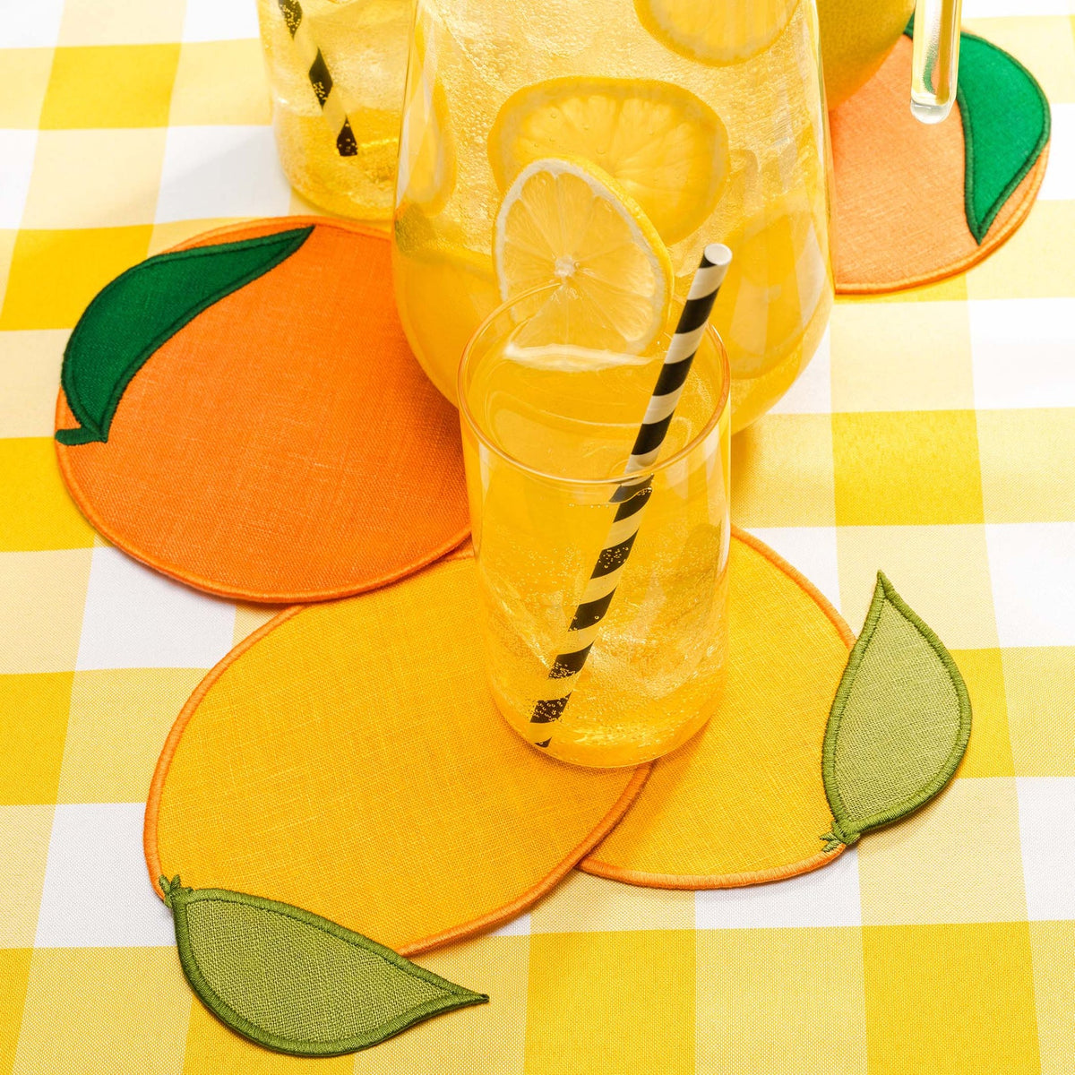 Round yellow and orange Piecework Citrus Cocktail Napkins on a yellow gingham tablecloth, set with a glass of lemonade and a pitcher.