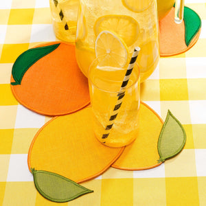Round yellow and orange Piecework Citrus Cocktail Napkins on a yellow gingham tablecloth, set with a glass of lemonade and a pitcher.