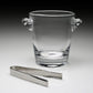 The William Yeoward Crystal Classic Ice Bucket, handmade with two handles, sits behind a pair of metal ice tongs on a neutral surface.