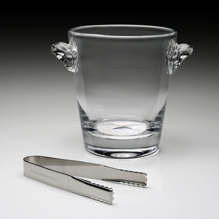 The William Yeoward Crystal Classic Ice Bucket, handmade with two handles, sits behind a pair of metal ice tongs on a neutral surface.