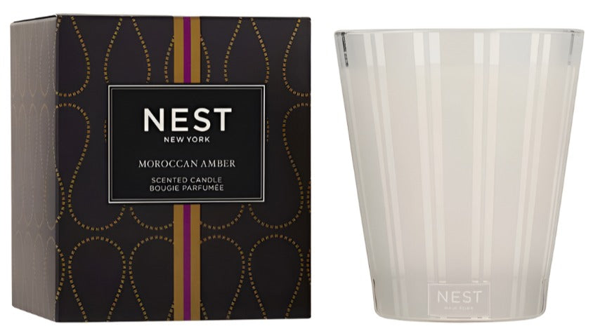 A Nest Moroccan Amber Candle from the Moroccan Amber Candle Collection is shown beside its black and gold box. The clear glass holder features subtle vertical stripes and contains a candle with patchouli notes.