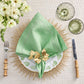 The Kim Seybert Classic Napkin Set, featuring an embroidered border, pairs elegantly with a gold napkin ring on a patterned plate and sunburst mat. Bamboo-handled fork and knife flank it, glasses are placed above, with white flowers to the left.