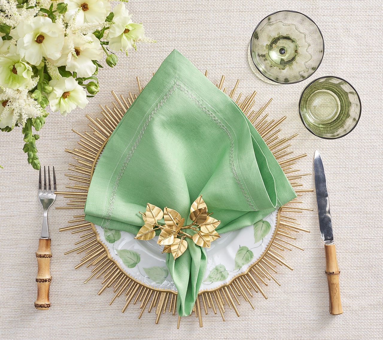 The Kim Seybert Classic Napkin Set, featuring an embroidered border, pairs elegantly with a gold napkin ring on a patterned plate and sunburst mat. Bamboo-handled fork and knife flank it, glasses are placed above, with white flowers to the left.