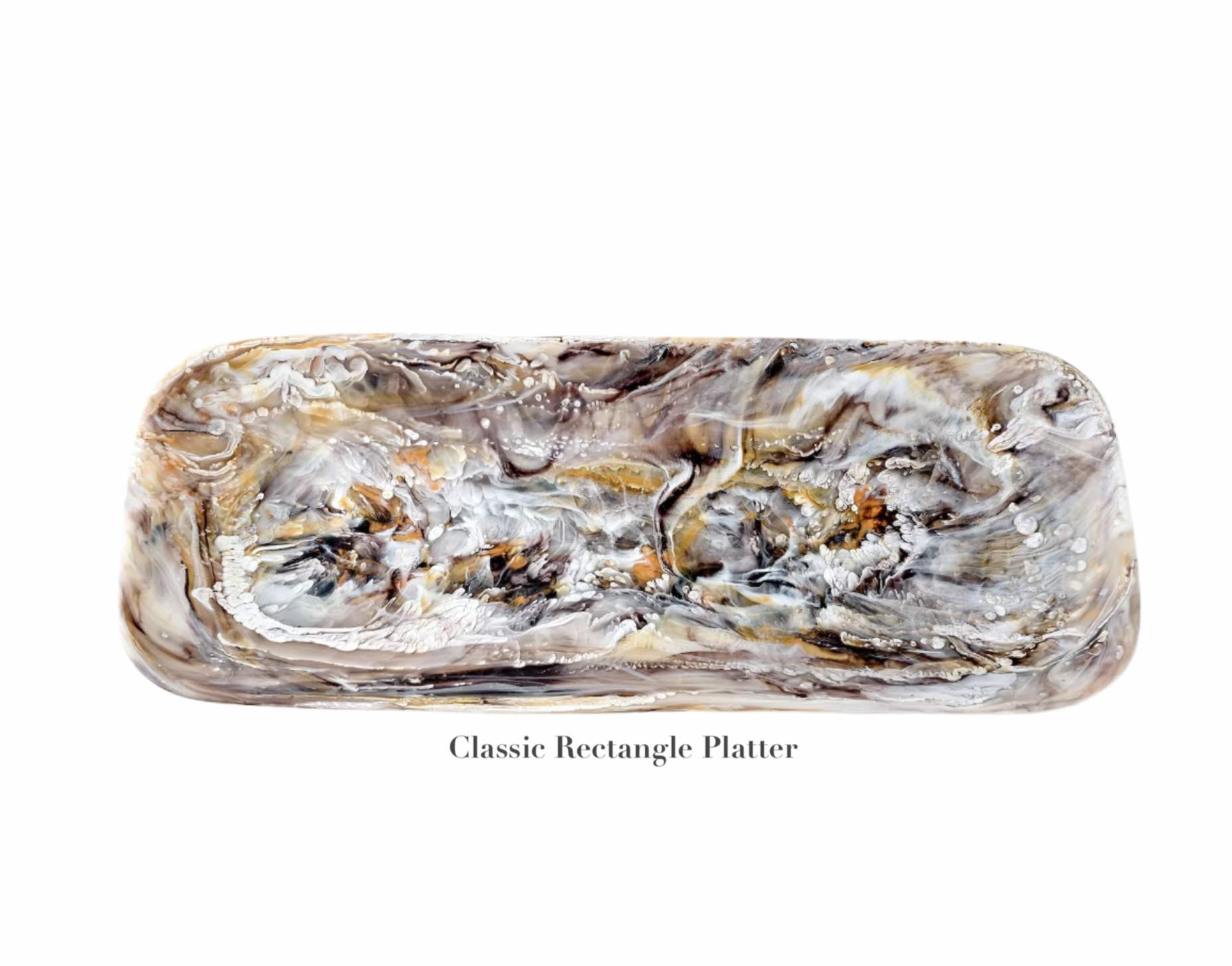 The "Mocha Resin Serveware Collection" by Nashi Home features a rectangular platter with a marbled gray, white, and brown design. Food safe and stylish, it’s perfect for home decor or serving guests.