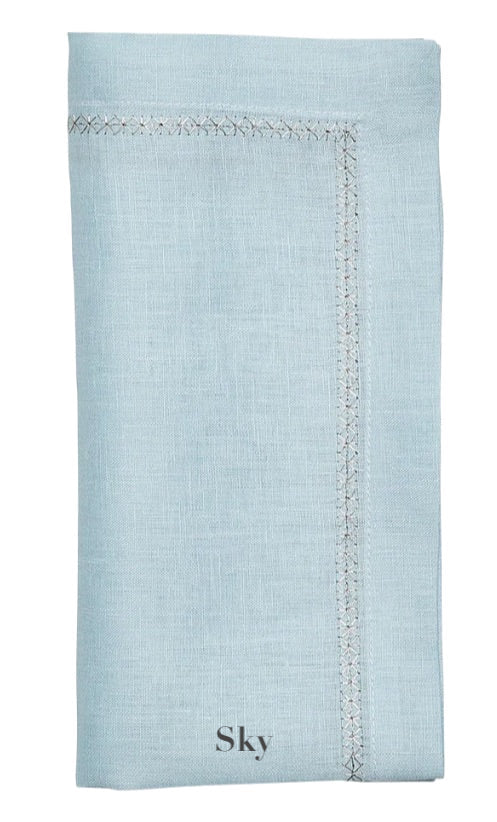 The Kim Seybert Classic Napkin Set features light blue linen napkins, elegantly labeled "Sky," with decorative stitching along the edges reminiscent of an embroidered border.