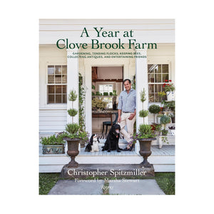 A man stands on a porch with two dogs, surrounded by potted plants at Clove Brook Farm. The book “A Year at Clove Brook Farm” by Common Ground captures the charm of this idyllic country retreat.