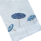 Haute Home’s Beach Umbrella Bath Collection features a white fabric napkin crafted from premium linen, adorned with three embroidered blue beach umbrellas arranged diagonally for a sophisticated touch.