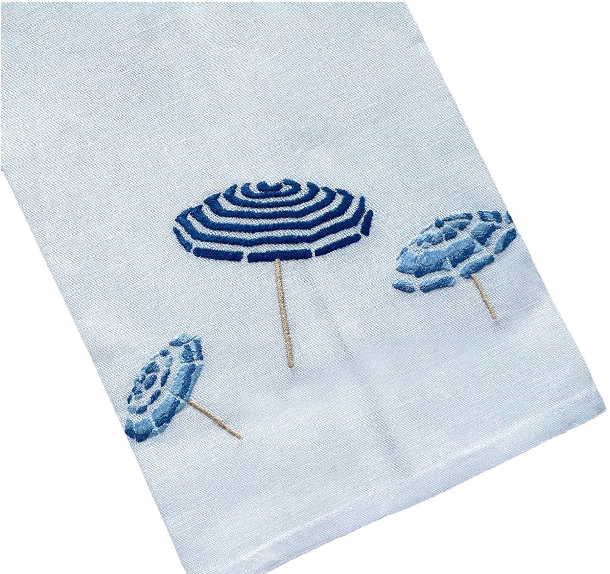 Haute Home’s Beach Umbrella Bath Collection features a white fabric napkin crafted from premium linen, adorned with three embroidered blue beach umbrellas arranged diagonally for a sophisticated touch.