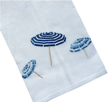 Haute Home’s Beach Umbrella Bath Collection features a white fabric napkin crafted from premium linen, adorned with three embroidered blue beach umbrellas arranged diagonally for a sophisticated touch.