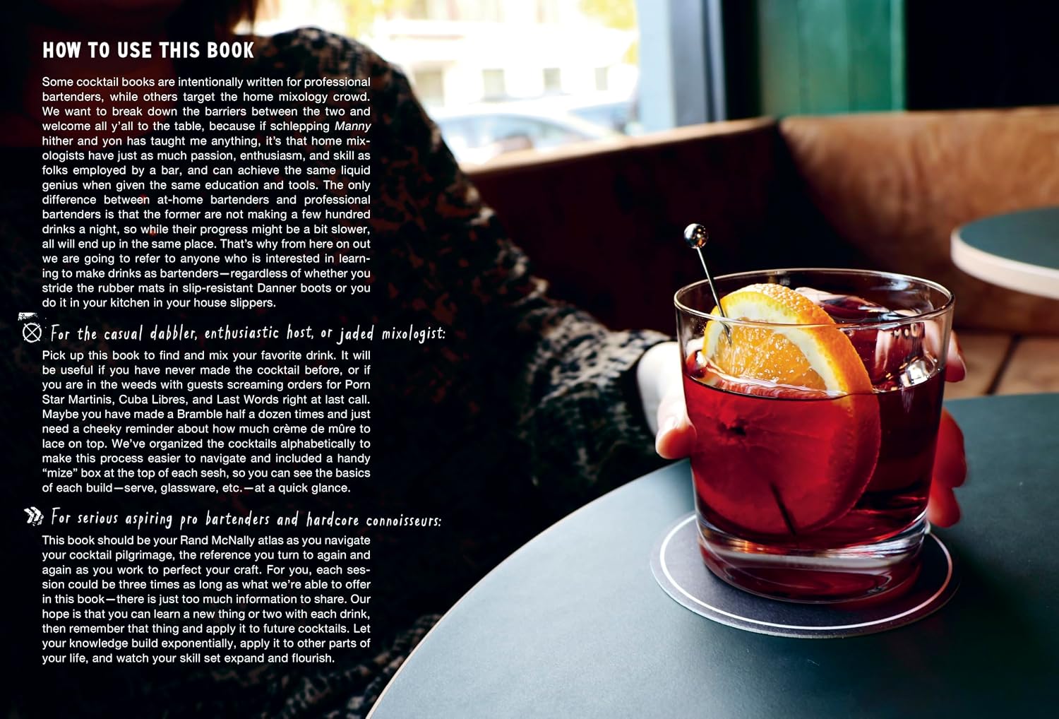 A close-up of a craft cocktail with an orange slice and ice sits in a glass on a coaster atop a table; nearby, a person with a patterned sleeve rests their arm. Instructions from The Classic Cocktail Sessions by Penguin Random House are visible, offering insight into mixology techniques.
