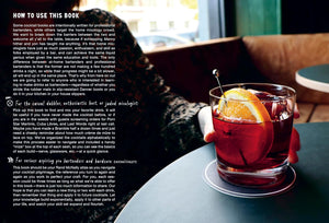 A close-up of a craft cocktail with an orange slice and ice sits in a glass on a coaster atop a table; nearby, a person with a patterned sleeve rests their arm. Instructions from The Classic Cocktail Sessions by Penguin Random House are visible, offering insight into mixology techniques.