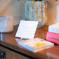 An Anne Neilson: Comfort Scripture Card featuring a Bible verse stands on a wooden table near a candle, a plant in a textured pot, books, and a stack of coasters.