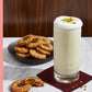 A tall glass of frothy white lassi, evoking memories of nostalgic cocktails, is topped with chopped pistachios and rests on a napkin beside a plate of cookies and a copy of Cookies & Cocktails by Simon & Schuster on a marble table.