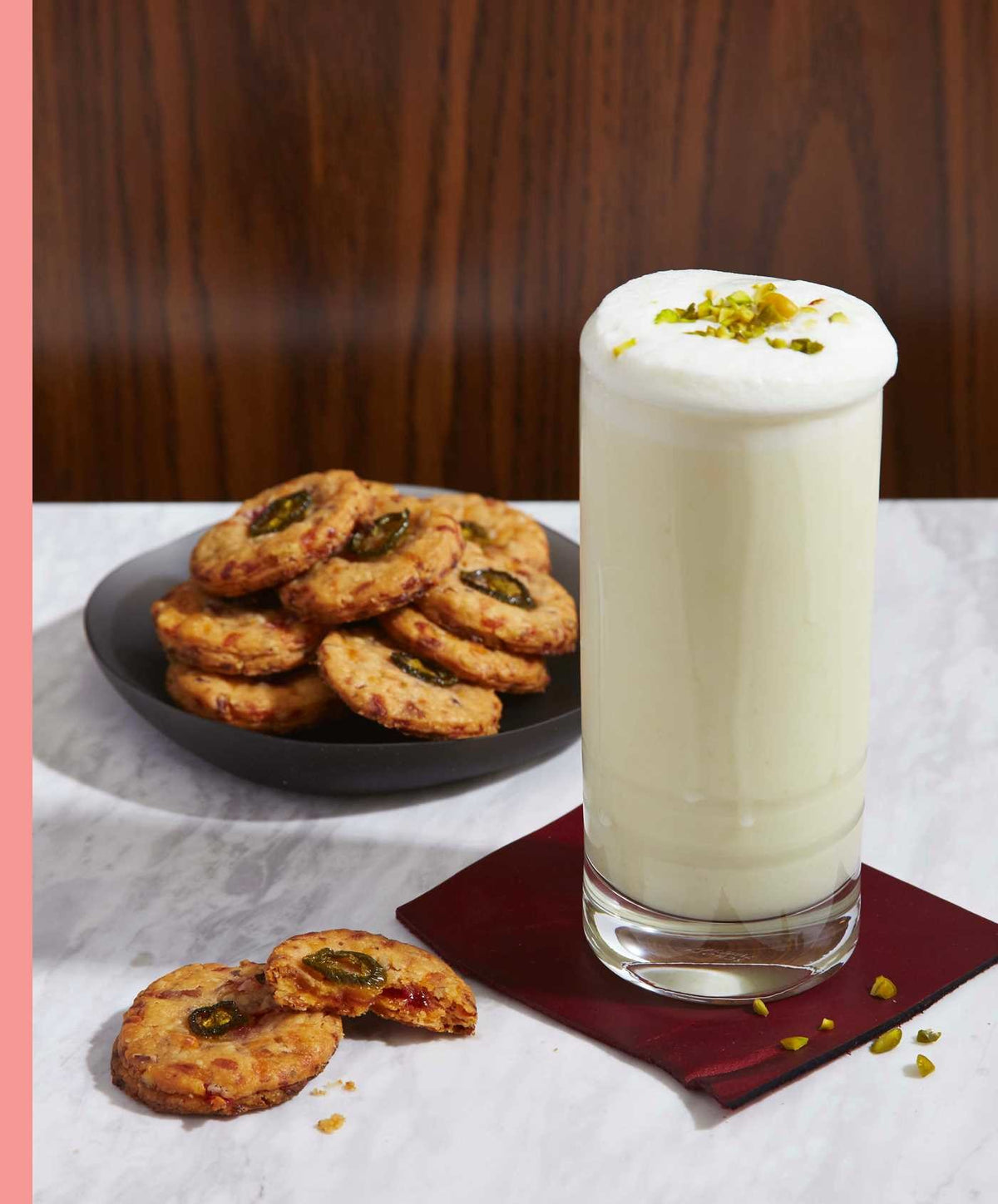 A tall glass of frothy white lassi, evoking memories of nostalgic cocktails, is topped with chopped pistachios and rests on a napkin beside a plate of cookies and a copy of Cookies & Cocktails by Simon & Schuster on a marble table.