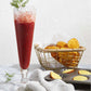 A tall glass of red iced Cookies & Cocktails by Simon & Schuster, garnished with dill, sits on a marble surface next to a basket of waffle-cut chips and a towel holding a baking tray filled with round crackers.