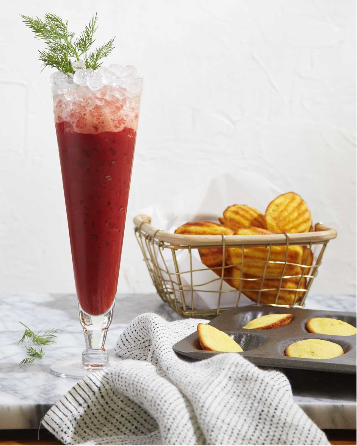 A tall glass of red iced Cookies & Cocktails by Simon & Schuster, garnished with dill, sits on a marble surface next to a basket of waffle-cut chips and a towel holding a baking tray filled with round crackers.
