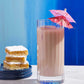 A tall glass of chocolate milk adorned with a pink paper umbrella, evoking nostalgic cocktails, is placed beside three powdered sugar lemon bars stacked on a dark plate—the perfect pairing to enjoy with *Cookies & Cocktails* from Simon & Schuster.