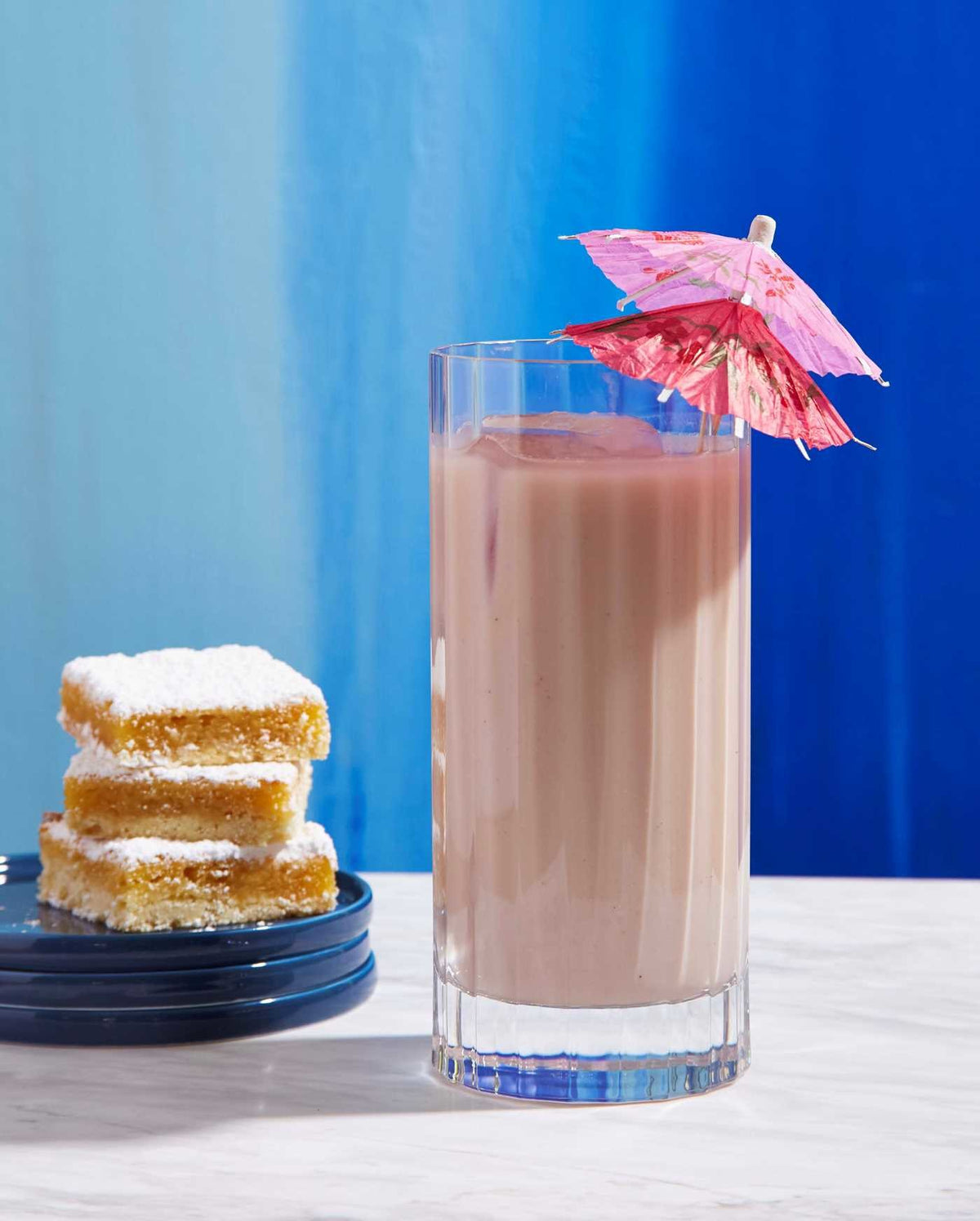 A tall glass of chocolate milk adorned with a pink paper umbrella, evoking nostalgic cocktails, is placed beside three powdered sugar lemon bars stacked on a dark plate—the perfect pairing to enjoy with *Cookies & Cocktails* from Simon & Schuster.