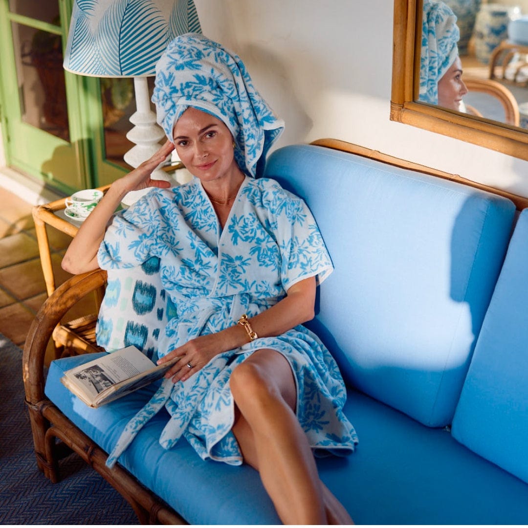 In a chic Palm Beach setting, a person in a scallop-trimmed blue and white robe from the Weezie Towels x Meg Braff Bath Collection lounges on a blue sofa with an open book, while their serene image is reflected in a mirror.