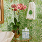 A bathroom corner showcases Weezie Towels x Meg Braff Cornflower Bath Accessories, featuring a tissue box with the Madame Butterfly pattern and a green leaf towel, accentuating the pink orchids against the green floral wallpaper.
