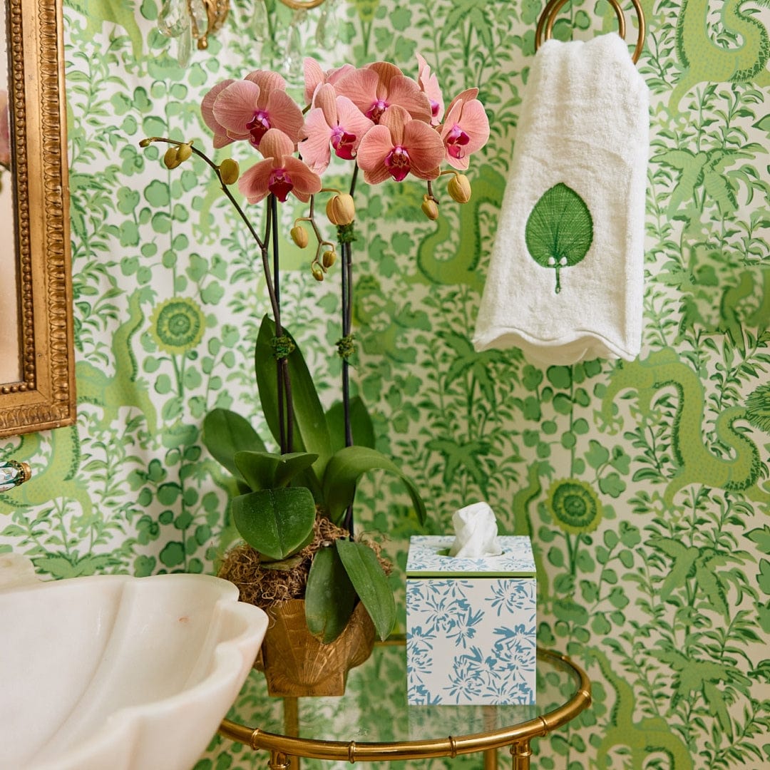 A bathroom corner showcases Weezie Towels x Meg Braff Cornflower Bath Accessories, featuring a tissue box with the Madame Butterfly pattern and a green leaf towel, accentuating the pink orchids against the green floral wallpaper.