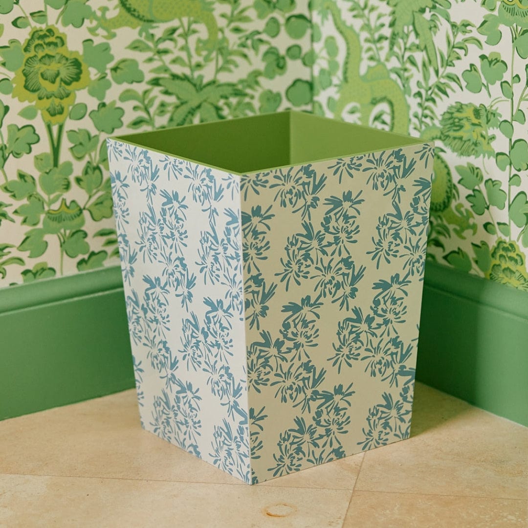 A Weezie Towels x Meg Braff Cornflower waste bin, featuring blue floral patterns reminiscent of Palm Beach charm, complements a room with green floral wallpaper and matching baseboard on a tiled floor.