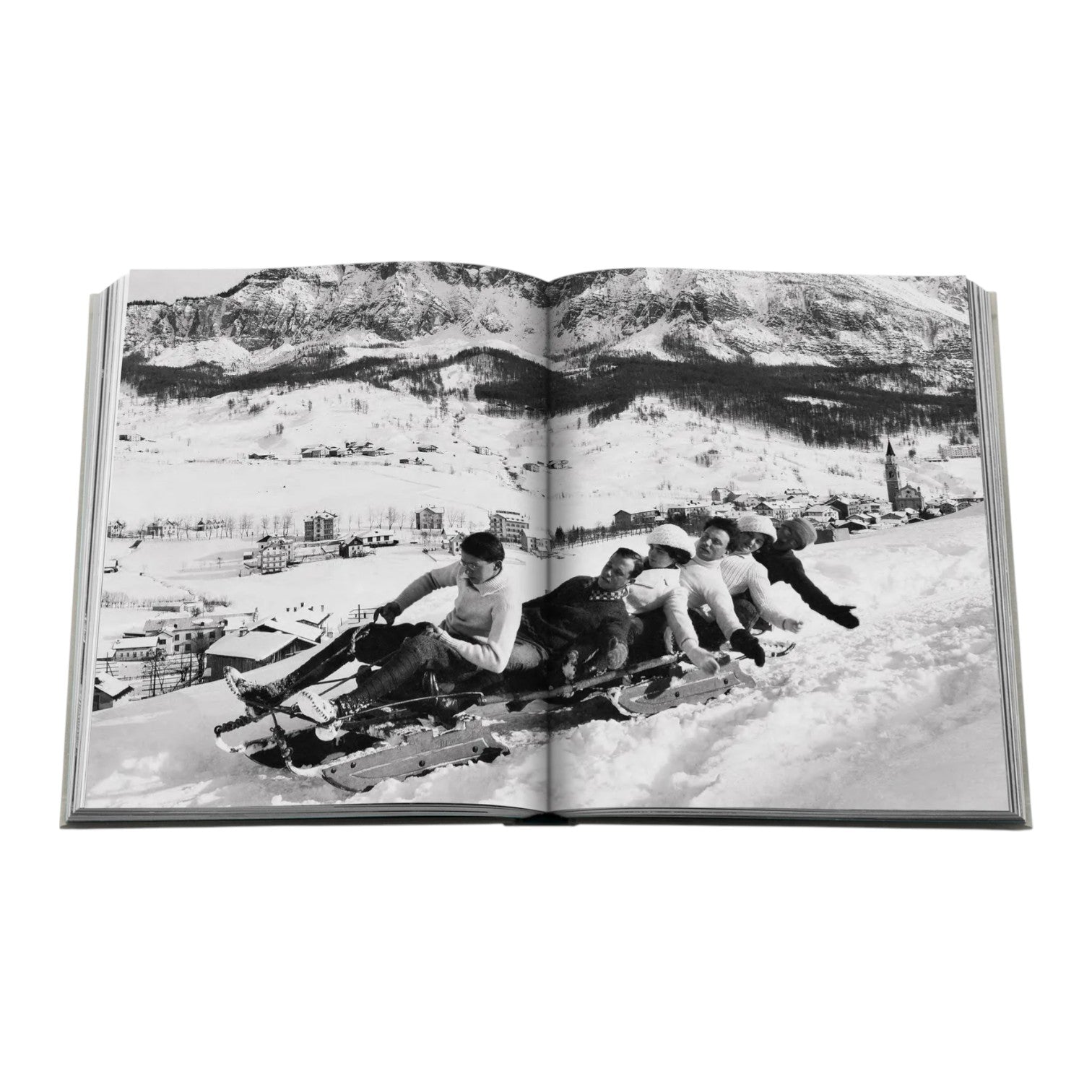 An open Assouline "Cortina d'Ampezzo" book shows a black-and-white photo of four people sledding on snowy hills near a mountain village, with the Dolomites rising in the background.
