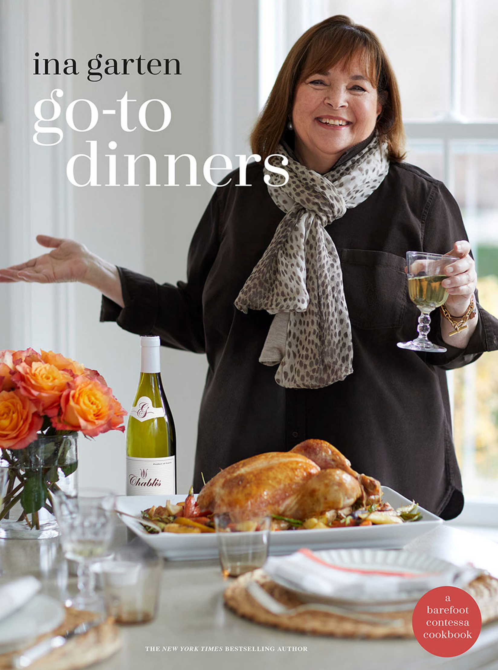 Ina Garten stands behind a set table with a roast chicken, holding a glass of white wine—a perfect showcase of comforting recipes from her book, “Go-to Dinners” by Penguin Random House. A bottle of wine and a bouquet of orange flowers are also on the table, creating an inviting scene inspired by Ina Garten’s delicious meals.