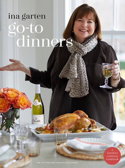 Ina Garten stands behind a set table with a roast chicken, holding a glass of white wine—a perfect showcase of comforting recipes from her book, “Go-to Dinners” by Penguin Random House. A bottle of wine and a bouquet of orange flowers are also on the table, creating an inviting scene inspired by Ina Garten’s delicious meals.