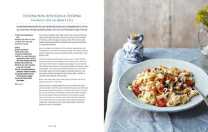A bowl of pasta with squid, chickpeas, and cherry tomatoes rests on a blue plate beside a blue cloth napkin and a small white vase filled with yellow flowers. Inspired by the regional Italian recipes featured in Cucina Di Amalfi By Ursula Ferrigno from Simon & Schuster, this dish captures the flavors of the Amalfi Coast. Recipe text appears on the left.